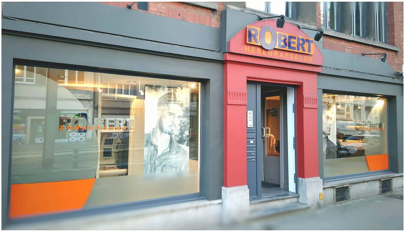 De gevel van Kapsalon Robert aan de Koning Albertlaan: rood portaal, de ROBERT-lichtbak en oranje cirkelvinyl op de etalages