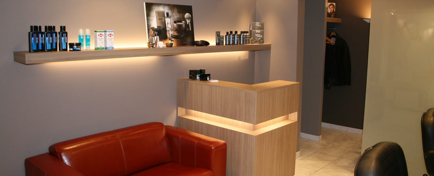 De receptiebalie van Kapsalon Robert met verlichte wandplank vol verzorgingsproducten en de rode leren zetel