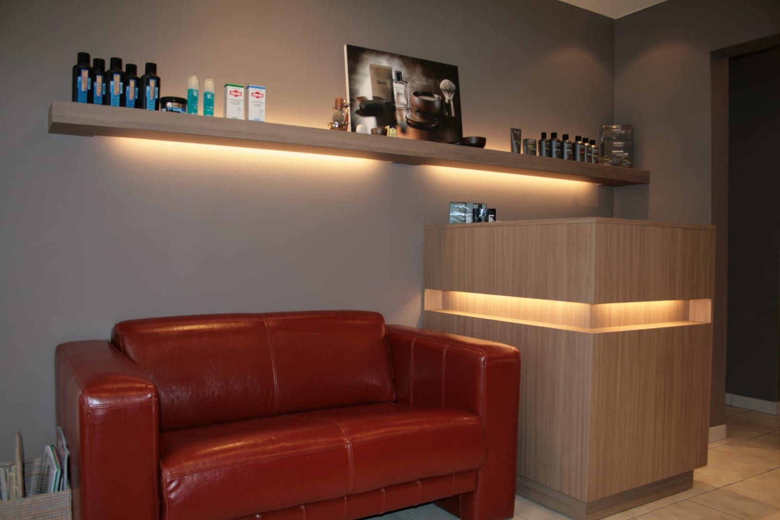 Het interieur van Kapsalon Robert met verlichte wandplank vol producten, de houten balie en de rode leren zetel